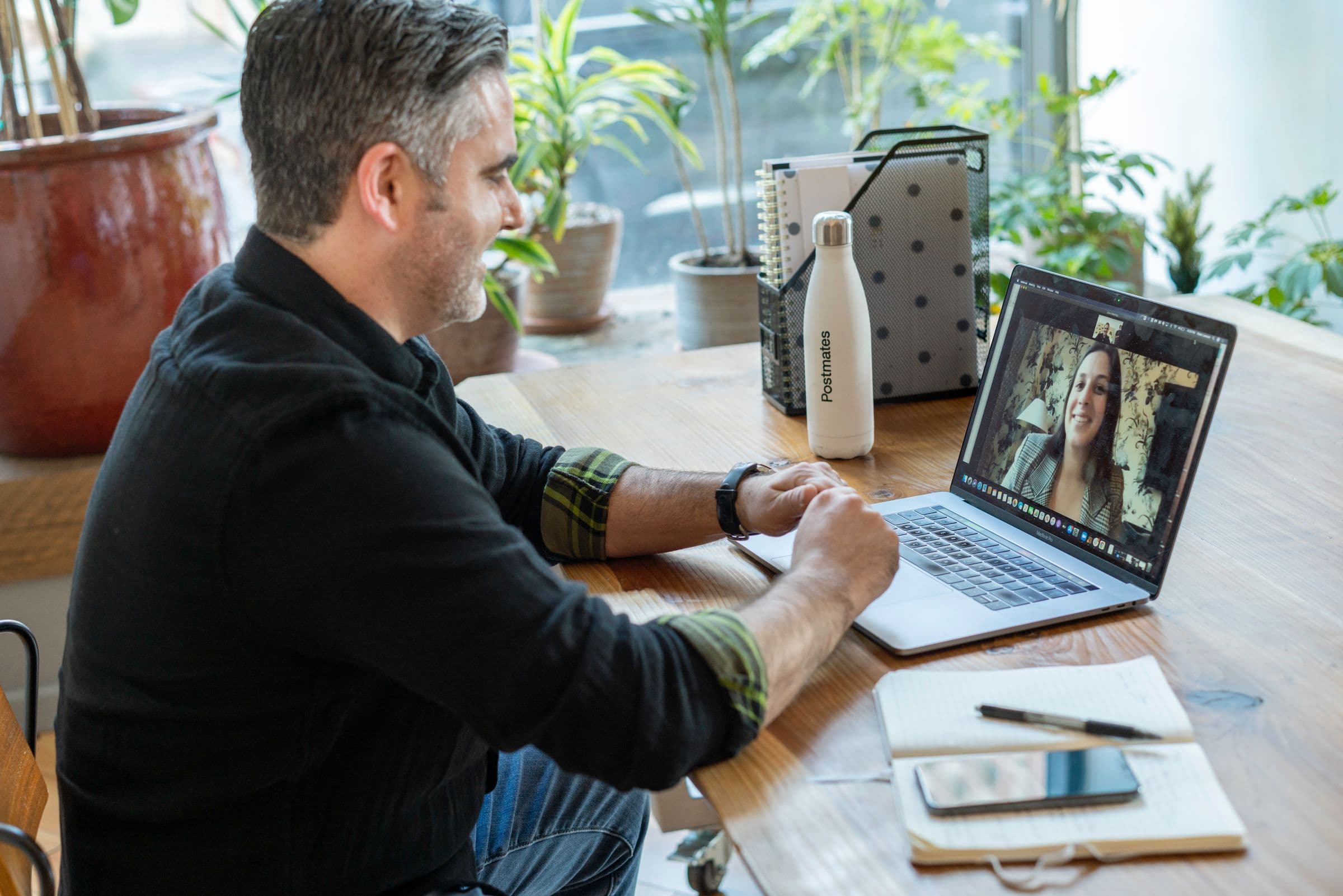 A man smiling on a video call at a wooden desk by the window, his laptop showing the colleague he is talking to